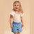 A young girl with blonde hair smiling, wearing a cream short-sleeve top with a tiny green and pink floral print and light blue denim shorts, set against a solid beige background.