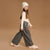 A young girl wearing a cream knit beanie, cream long-sleeve top, dark gray wide-leg pants with a small white floral pattern, and cream chunky platform shoes, walking against a solid tan studio background.
