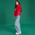 A young girl modeling a bright red plush fleece sweatshirt with a colorful graphic on the chest, paired with light-wash wide-leg jeans and white sneakers, set against a solid green studio background.