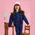 A young girl with long brown hair wearing a navy blue and white polka dot matching set, consisting of a long-sleeve top with ruffle details and matching pants, posing in front of a pink background with wooden chairs and a globe.