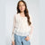 A young girl with long brown hair wearing a cream-colored long-sleeve blouse with sheer polka dot sleeves and a ruffled tiered bodice, paired with light blue jeans, against a light gray studio background.