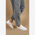 White low-top canvas sneakers with silver eyelets and a thin red stripe, shown on a model wearing gray jeans against a solid tan background.