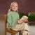 A young girl with blonde hair in a ponytail sits on a wooden chair, wearing a light green knit sweater and beige trousers while holding a rolled parchment map. The background features a textured dark plum wall, a potted green plant, and a small red-and-white mushroom decoration.