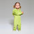 A toddler girl with blonde pigtails stands in a studio wearing a bright lime green hooded one-piece pajama or onesie with fluffy ear details on the hood. She is posed against a neutral light gray background.