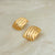 A pair of gold-toned textured stud earrings with a ribbed, wave-like pattern, displayed on a light beige marble surface.