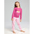 A young girl stands barefoot on a white textured rug against a light gray studio background, wearing a pink long-sleeve pajama top with a crown graphic and matching pink patterned pajama pants.
