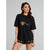 A woman modeling an oversized black t-shirt with a gold bee graphic and the text 'Jamboree' on the chest, paired with black shorts featuring a gold bee pattern, against a solid gray studio background.