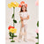 A young girl stands in a studio wearing a white short-sleeve top, cream pants with small blue floral prints, white sneakers, and holding a cream crescent-shaped handbag. She is wearing a large coral-orange flower headband.