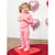 A young girl with blonde pigtails stands smiling on a red heart-shaped platform, wearing a pink long-sleeve pajama set with a subtle heart pattern. The background features a white wall with pink heart-shaped balloons and a wooden floor.