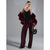 A model stands against a gray studio background wearing a black camisole, wide-leg sequined trousers in deep burgundy, a matching burgundy faux-fur jacket draped off the shoulders, and holding a black handbag with gold hardware.