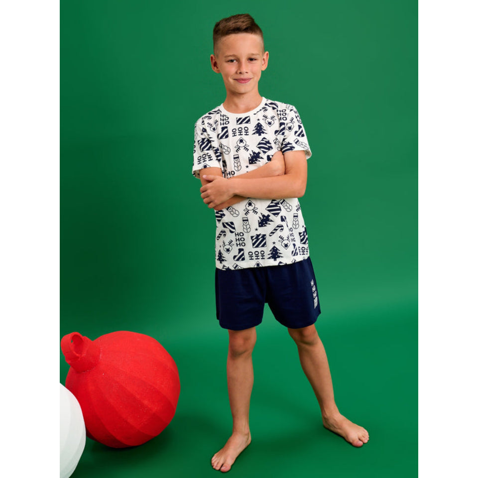 A young boy stands barefoot against a solid green studio background, wearing a white short-sleeve t-shirt with a navy blue graphic pattern and navy blue shorts. A large red ball prop is visible on the left side of the frame.