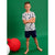 A young boy stands barefoot against a solid green studio background, wearing a white short-sleeve t-shirt with a navy blue graphic pattern and navy blue shorts. A large red ball prop is visible on the left side of the frame.