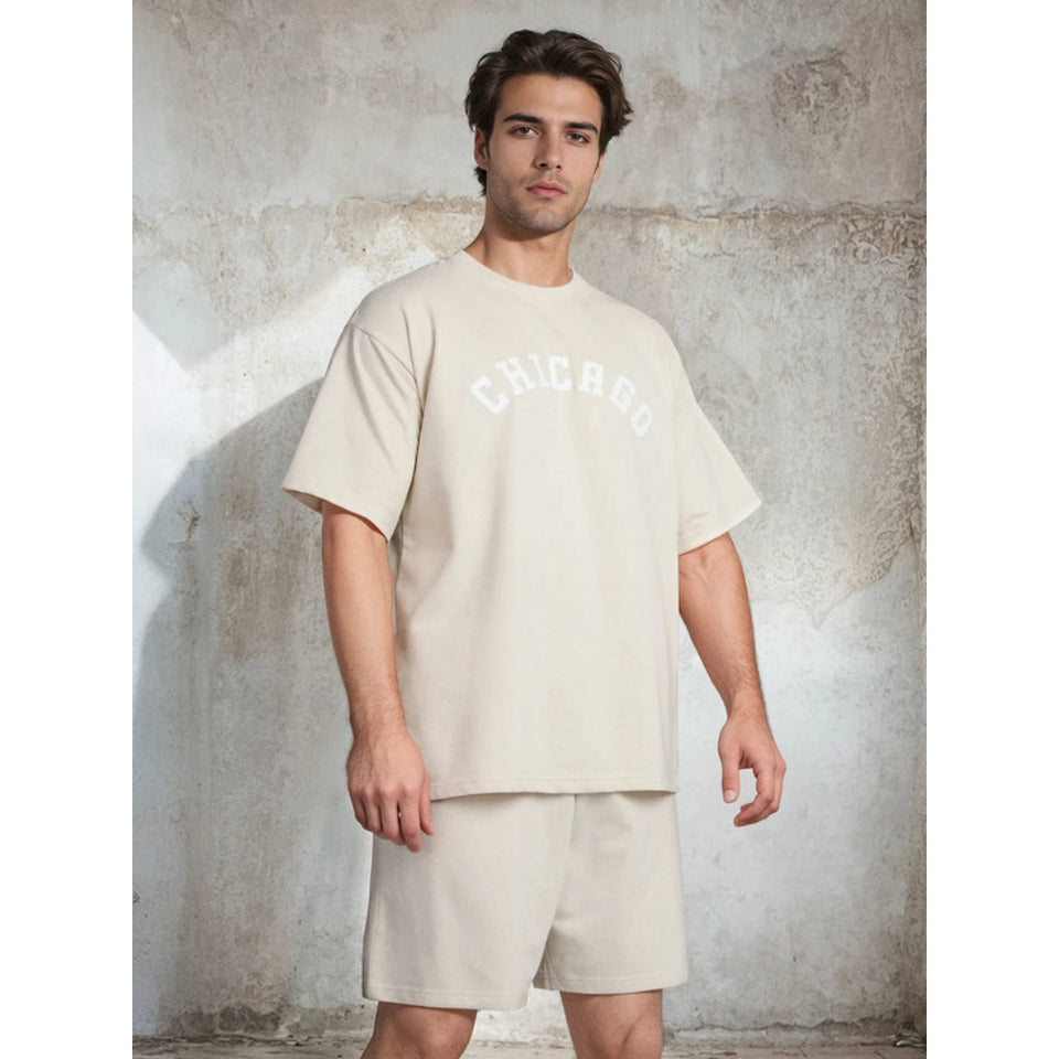 A man modeling a matching beige short-sleeve t-shirt and shorts set with 'CHICAGO' printed in white arched text across the chest, posed against a textured concrete wall.
