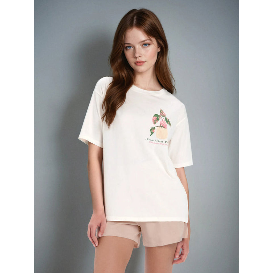 A woman modeling a cream-colored oversized t-shirt with a small pink floral graphic on the chest, paired with beige shorts, against a solid gray studio background.