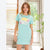 A young woman wearing a light aqua blue t-shirt dress with a retro 'Vacation Wood' graphic on the front, standing in a brightly lit room with a window and plant.