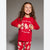 A young girl with long brown hair smiling and pointing at a red long-sleeve pajama top with gingerbread-themed graphics and the text 'Leave the cookies for Santa Claus'. She is wearing matching red pajama pants with gingerbread prints, set against a plain gray studio background.