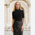 A woman stands in front of a stone archway wearing a fitted black short-sleeve mock-neck top and a black leather skirt with a studded belt.