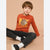 A young boy sitting on a beige studio floor wearing a rust-orange long-sleeve graphic t-shirt with a cartoon beaver and the text 'Beaver Jazz', paired with black jeans, white socks, and black sneakers.