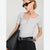 A woman wearing a light gray short-sleeve henley-style top, black pants, a black belt, black sunglasses, and layered necklaces, holding a black textured shoulder bag against a light gray studio background.