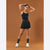 A woman modeling a black athletic tank top and matching pleated tennis skirt against a solid warm brown studio background.