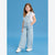 A young girl modeling light blue denim overalls with a white heart pattern, worn over a white t-shirt and white sneakers, set against a solid blue studio background.