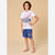 A young boy stands barefoot against a solid beige studio background, wearing a white graphic t-shirt with a skateboard design and blue shorts with a skateboard print on the leg.