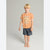 A young boy with blonde hair stands barefoot against a light gray studio background, wearing a short-sleeve orange graphic T-shirt with a dinosaur print and the words 'TYRANNO SAURUS' and dark gray shorts.