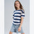 A young girl with long light brown hair is wearing a navy blue and white horizontally striped short-sleeve t-shirt and light-wash denim shorts, standing against a light gray studio background.