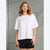 A woman modeling a white short-sleeve t-shirt with small rhinestone embellishments across the chest, paired with black shorts, standing against a textured concrete wall.