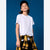 A child model wearing a plain white short-sleeve crewneck t-shirt, a black baseball cap, and a yellow and black plaid flannel shirt tied around the waist, set against a solid blue studio background.