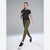 A woman modeling a black short-sleeve athletic top and olive green leggings with black sneakers, posed against a light gray studio background.