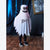 A child wearing a white ghost costume with black jagged hem and a spooky face on the hood, standing on a wooden floor in front of dark draped curtains.
