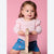 A toddler girl with light brown hair wearing a light pink long-sleeve ruffle-sleeve top and blue denim shorts with pink strawberry details, riding a small wooden toy on a pink background.