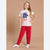A young girl modeling a two-piece pajama set consisting of a cream-colored t-shirt with a 'Winter Wonderland' graphic and red pants with a white heart pattern, set against a solid beige studio background.