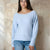 A woman wearing a light blue knit sweater with a wide boat neckline and long sleeves, paired with blue jeans, standing against a textured beige wall.