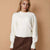 A woman wearing a cream-colored knit sweater and a brown skirt, standing against a solid beige background.