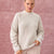A woman wearing a chunky knit cream-colored mock neck sweater, standing against a textured dusty pink background.
