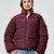 A woman wearing a cropped, quilted puffer jacket in a deep burgundy or maroon color, paired with blue jeans, against a light gray studio background.