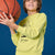 A smiling boy holding a basketball, wearing a light yellow-green long-sleeve sweatshirt with a black cartoon graphic and 'SOUTH VIEW' text, posed against a solid blue background.