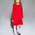A young girl modeling a bright red long-sleeve dress with ruffle details on the shoulders, paired with light pink tights and black ballet flats, against a neutral gray studio background.