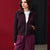A woman wearing a dark plum zip-up jacket over a white graphic T-shirt and matching plum pants, standing against a split red and black studio background.