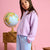 A young girl with long brown hair wearing a lavender waffle-knit sweater and light blue pants, posing next to a globe on a wooden stand against a solid pink background.