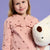 A young girl with blonde hair smiling while wearing a pink zip-up jacket with embroidered brown stars and holding a white plush toy against a solid lavender background.