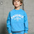 A young boy wearing a bright blue crewneck sweatshirt with a white 'PHOENIX SUPER 60 TEAM' graphic on the front, standing against a textured concrete wall.