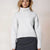 A woman wearing a light heather gray ribbed turtleneck sweater and a dark gray skirt, standing against a plain light beige studio background.