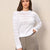 A woman wearing a white long-sleeve knit sweater with an openwork crochet pattern, paired with brown and white houndstooth trousers, standing against a textured beige wall.