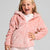 A young girl with blonde hair smiling and laughing while wearing a pink hooded zip-up fleece jacket over a white patterned shirt, set against a plain light gray studio background.