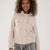 A woman wearing a light beige cropped button-up jacket with flap chest pockets, paired with gray drawstring pants, against a plain off-white studio background.