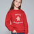A young girl with long brown hair wearing a red crewneck sweatshirt with 'Merry Sweetmas' and a gingerbread man graphic on the front, paired with blue jeans, against a solid gray studio background.