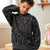 A child wearing a dark gray and black abstract patterned hoodie sweatshirt, standing in a softly lit room with a teddy bear visible in the background.